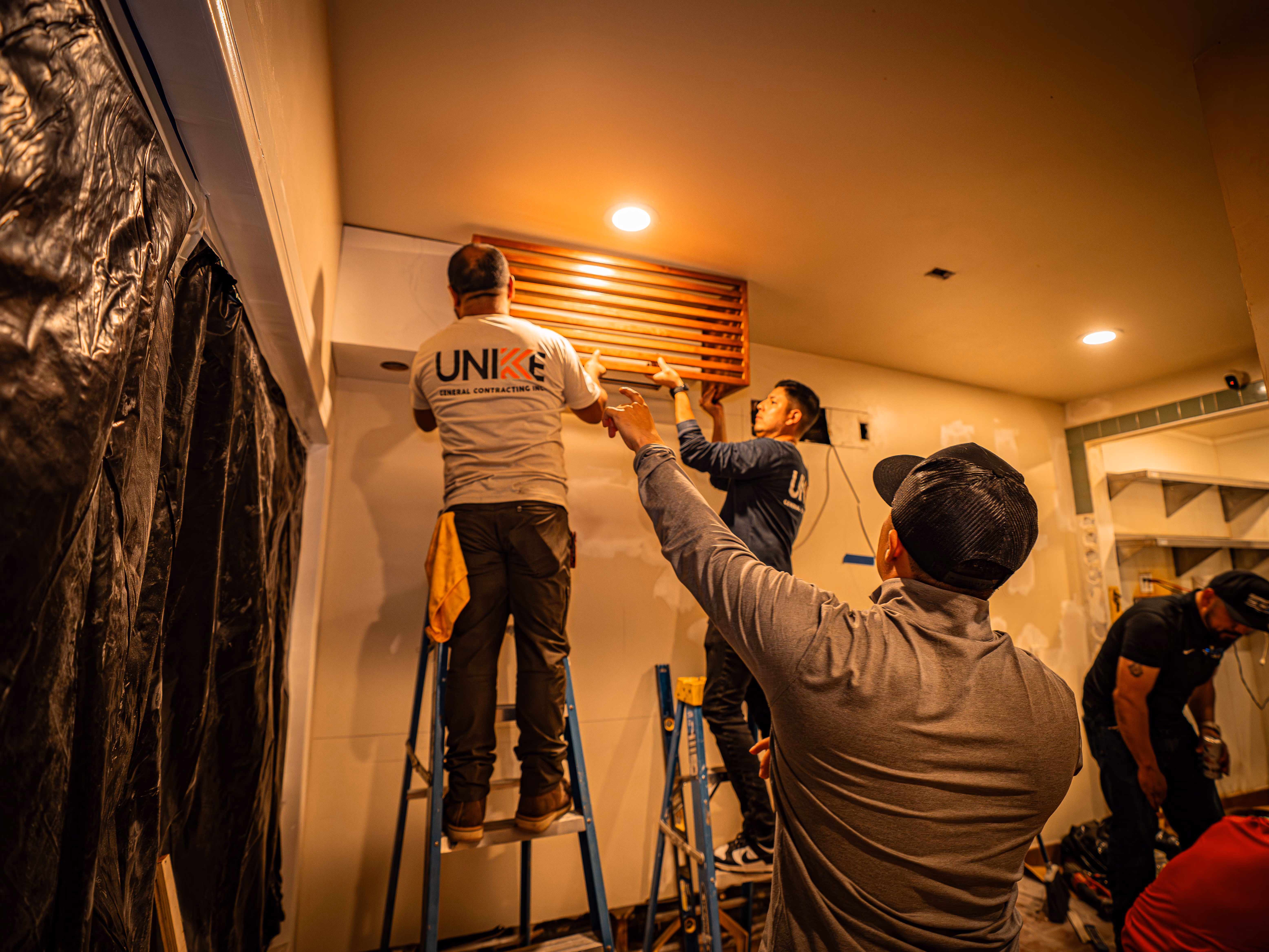 Three men installing a wooden vent cover on a wall using ladders in a construction area.