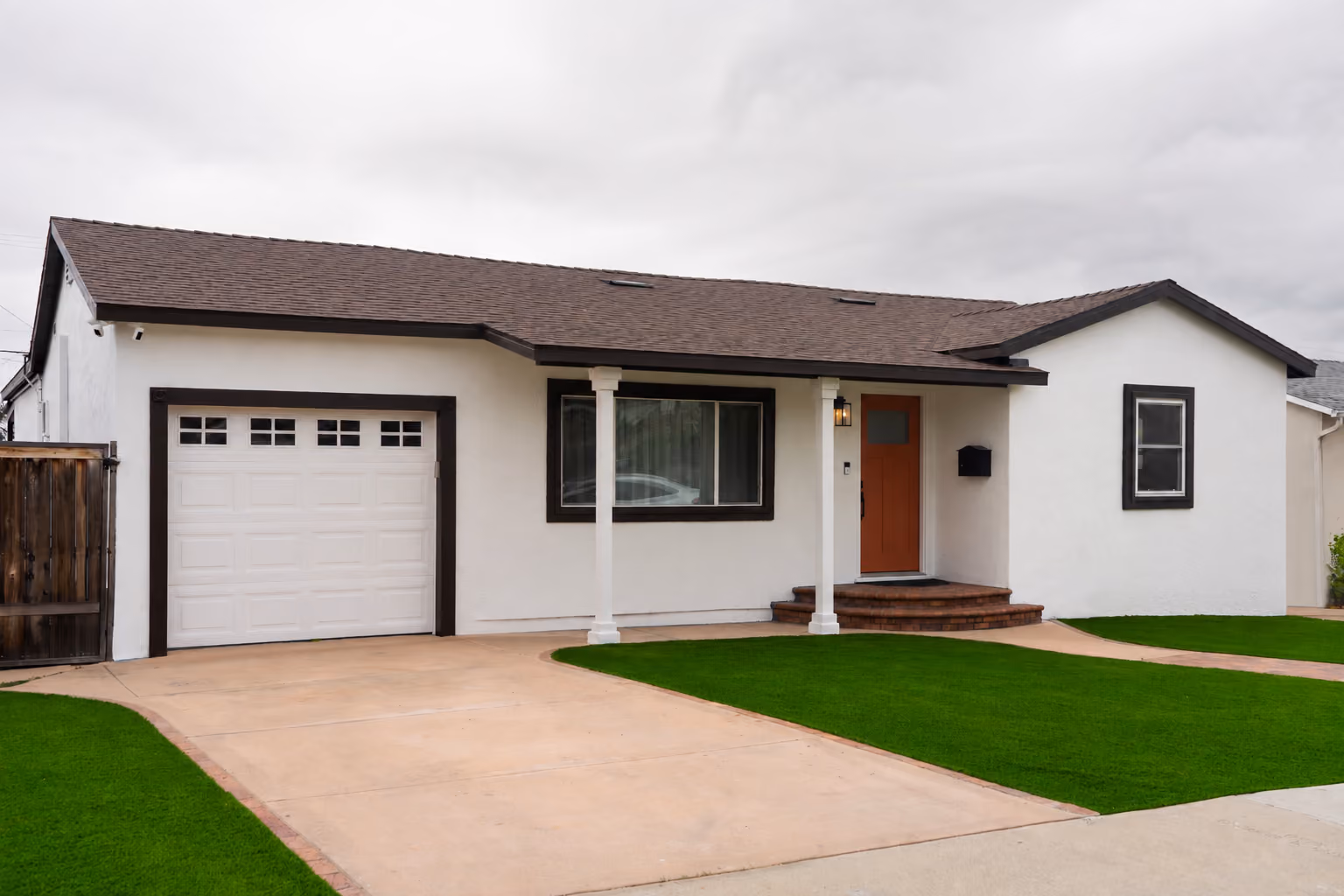 Single-story white house with brown roof, red front door, garage, and green lawn under gray sky.
