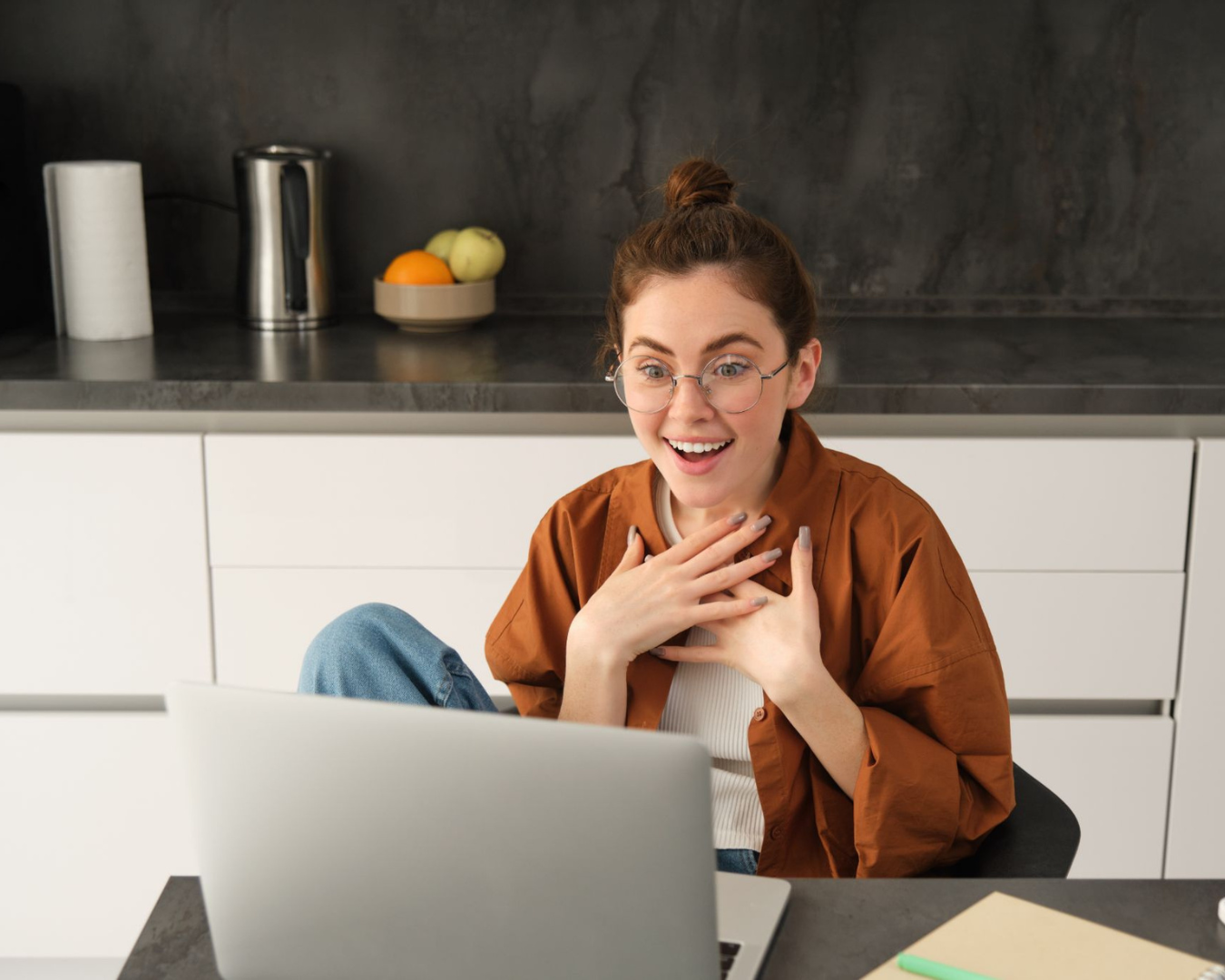 Excited young woman with glasses using laptop in modern kitchen, hands on chest in surprise.