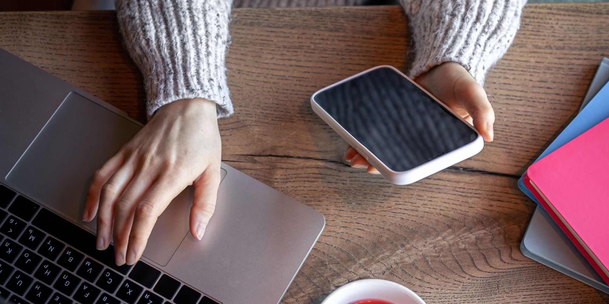 Person's hands using a laptop and holding a smartphone on a wooden desk with notebooks.
