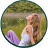 Woman in white top and lavender pants sitting on grass by a lake with eyes closed, head back