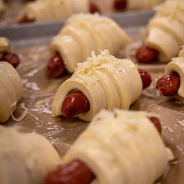 Uncooked pigs in a blanket with sausages wrapped in dough topped with shredded cheese on baking tray.