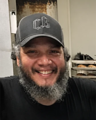 Smiling man with beard wearing a cap and black shirt in a bakery with bread on racks behind him.