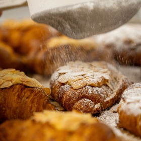 Powdered sugar being sprinkled onto almond croissants arranged on a tray.