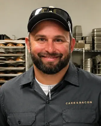 Smiling man with a beard wearing a Cake & Bacon shirt and a black cap in a bakery kitchen.