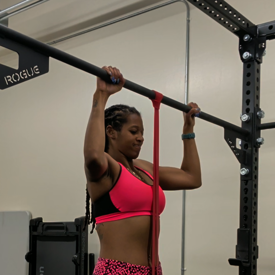 Woman in a pink sports bra using a red resistance band on a pull-up bar in a gym.