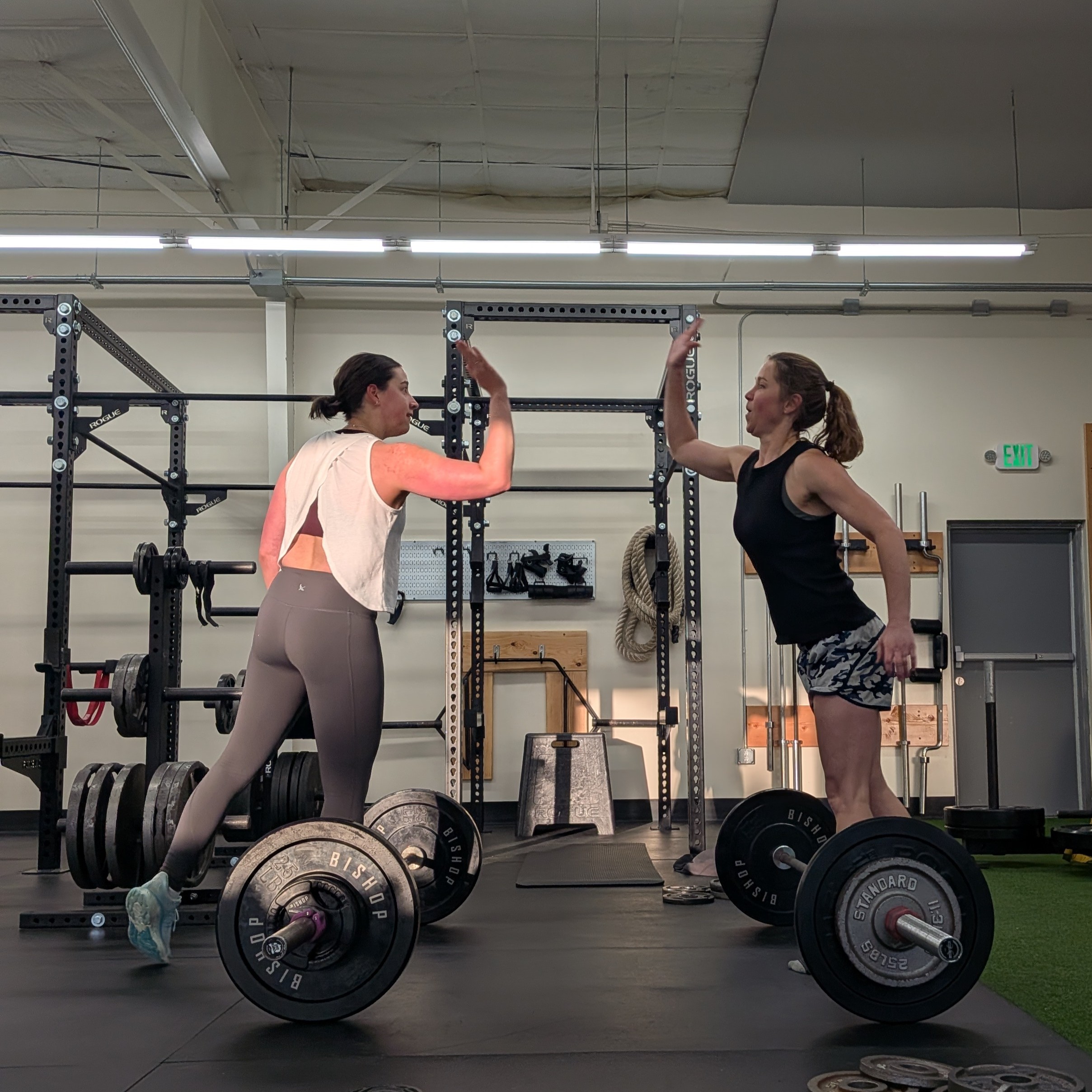 Two women in a gym giving a high five while standing near barbells on the floor.