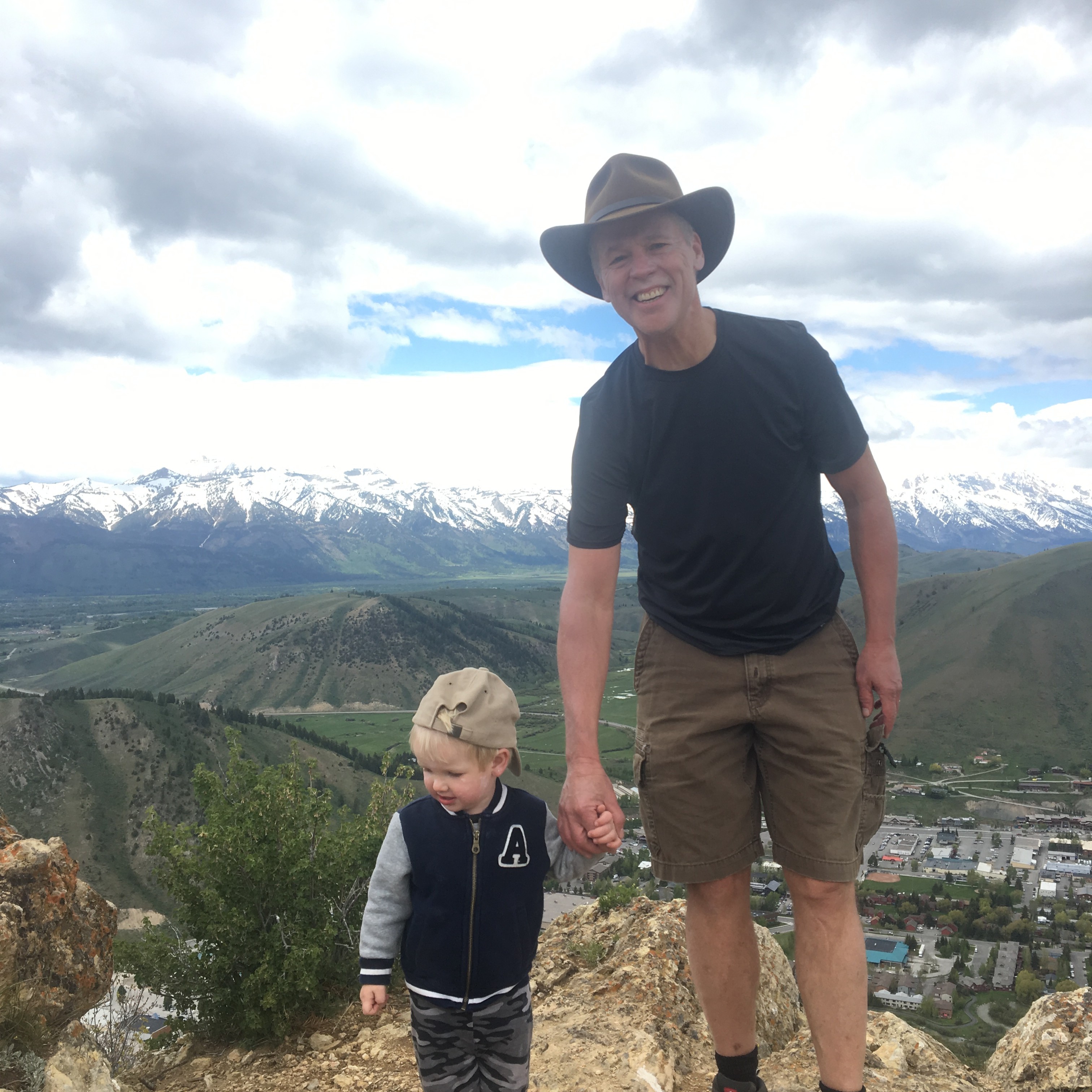 Man wearing a hat holding hands with a young child on a rocky hill with mountains in the background.