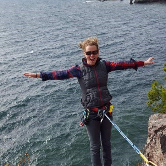 Woman in sunglasses and climbing gear standing on a rocky cliff edge above a large body of water.