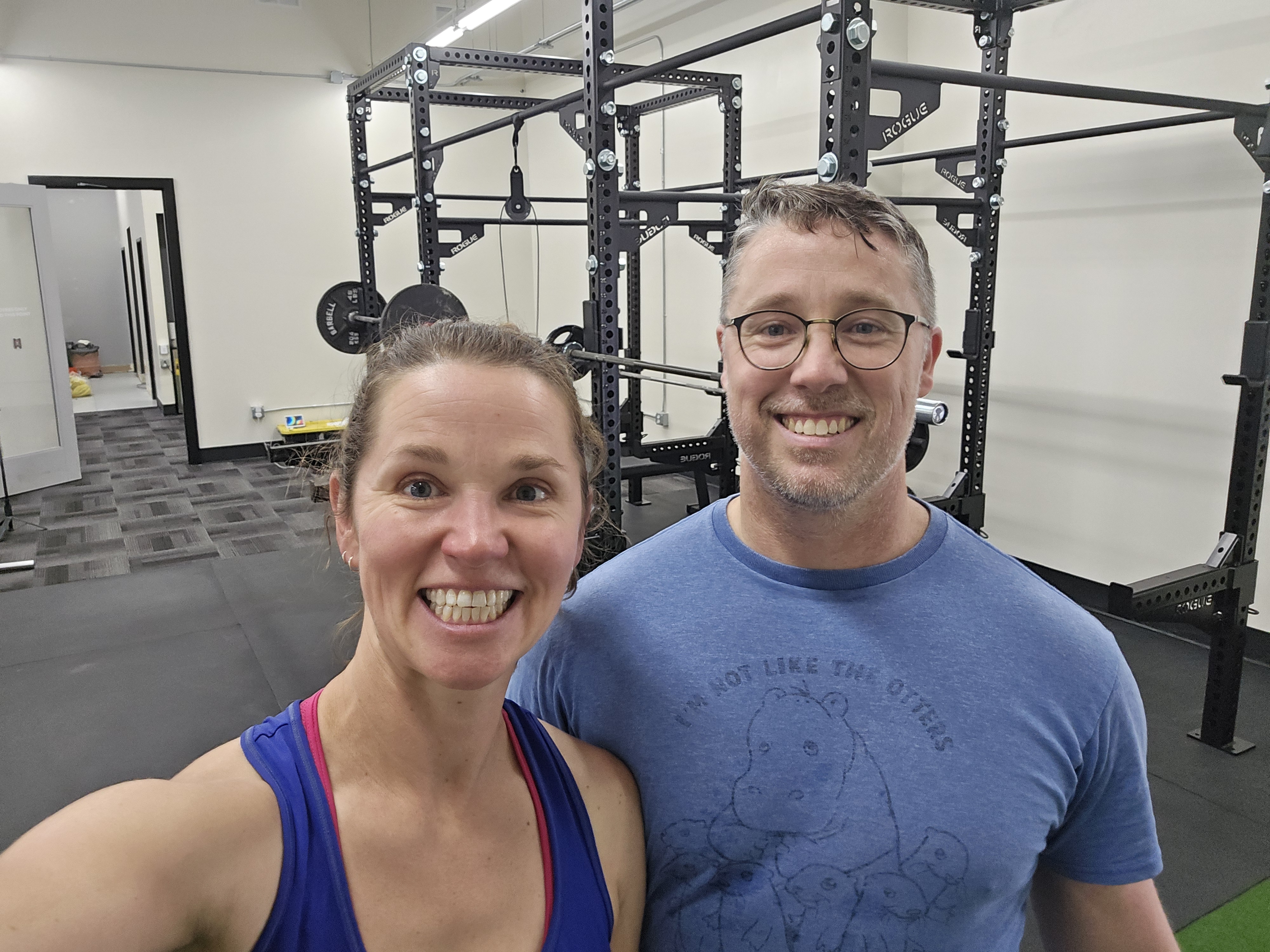 Smiling man and woman posing for a selfie in a gym with weightlifting racks behind them.