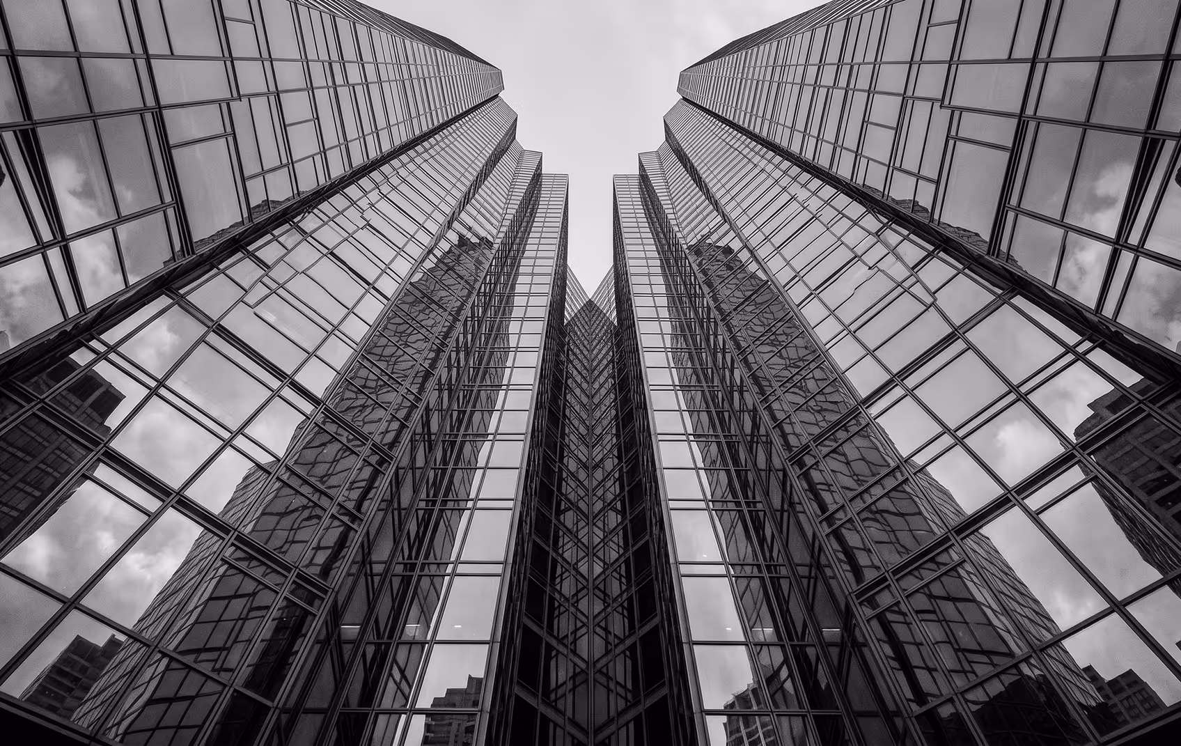 Black and white photo looking up at glass skyscrapers reflecting surrounding buildings and cloudy sky.