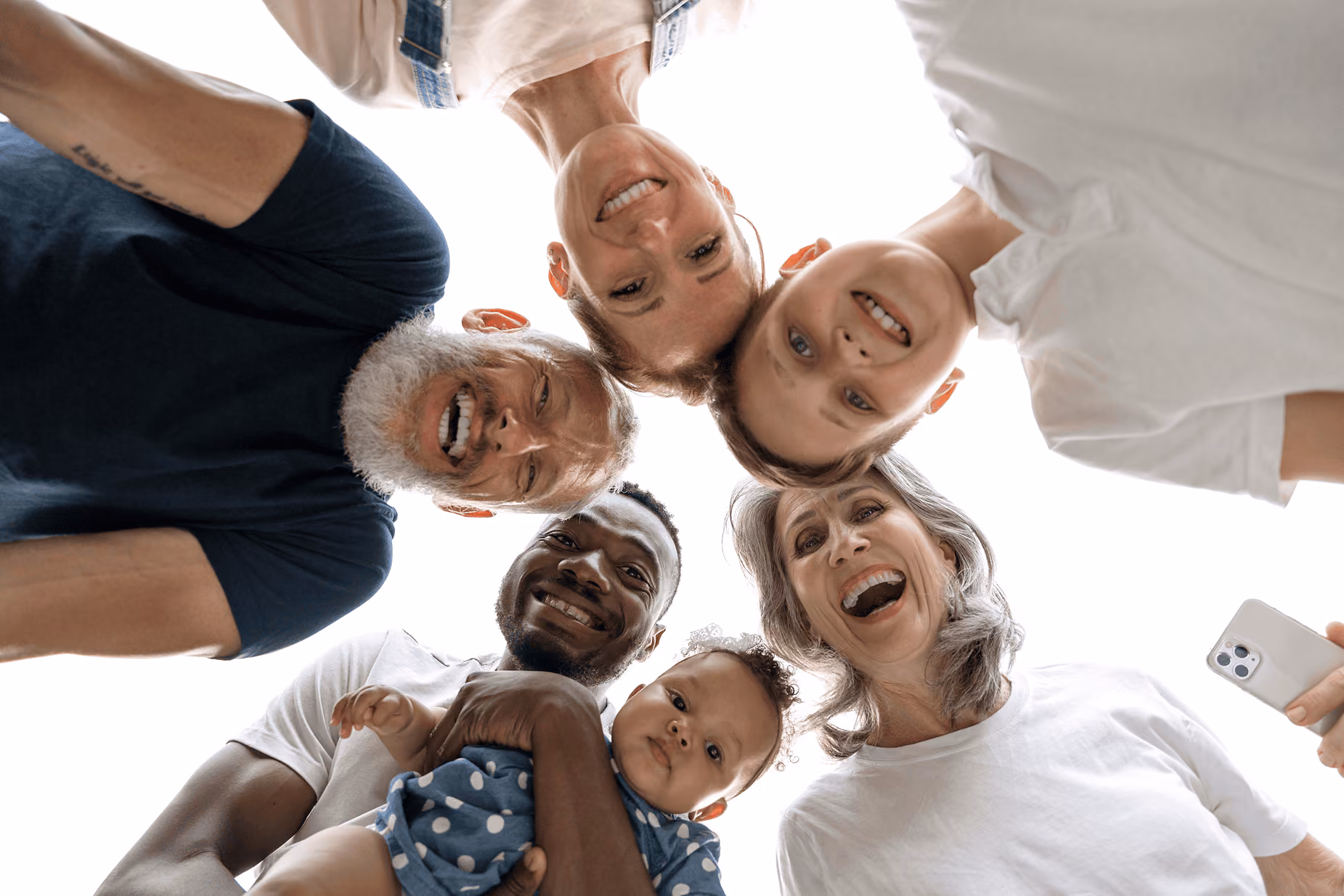 Multigenerational diverse family smiling in a circle looking down at the camera, holding a baby.