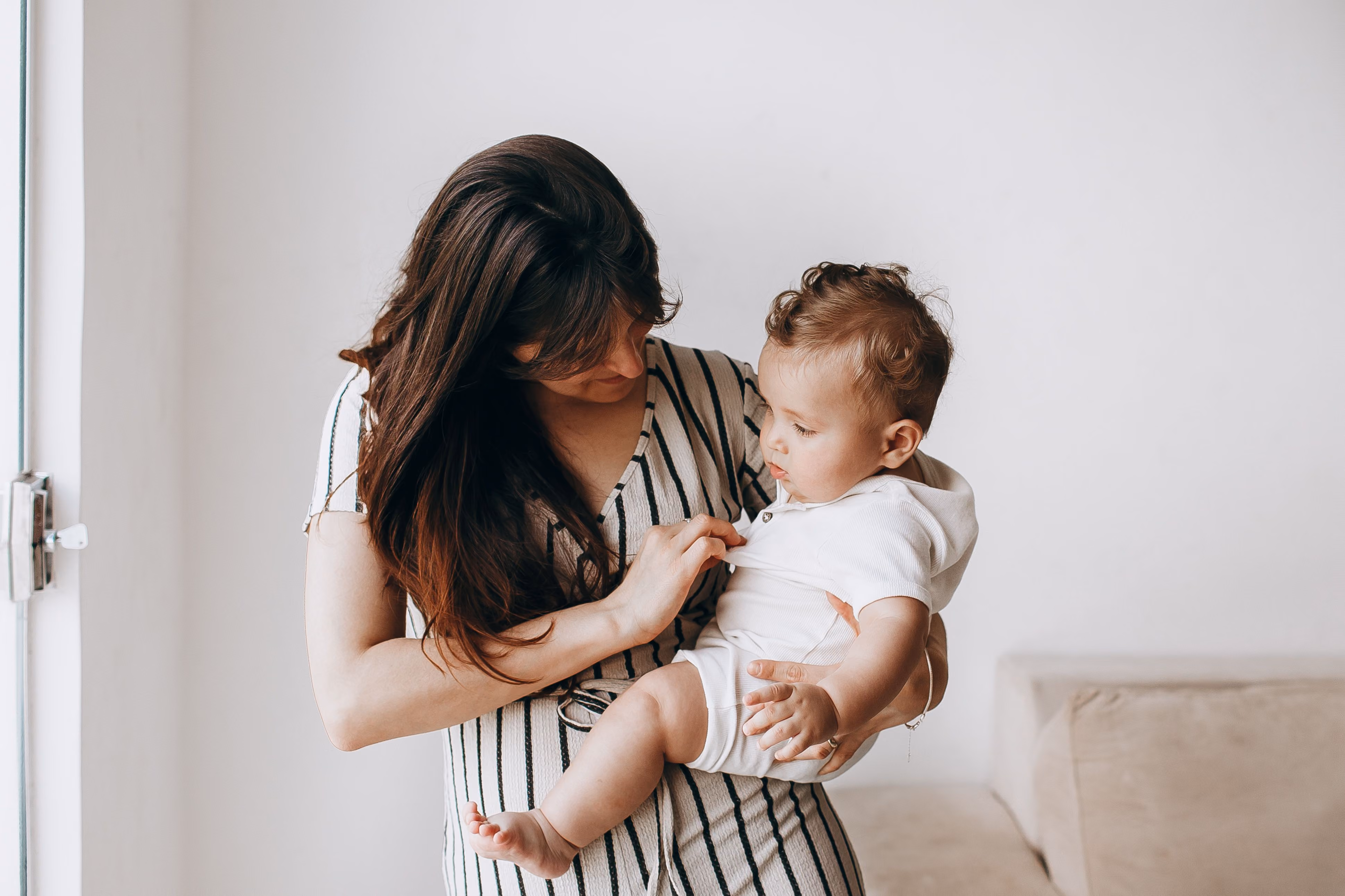 Woman in striped dress holding a baby wearing a white outfit near a beige couch and window.