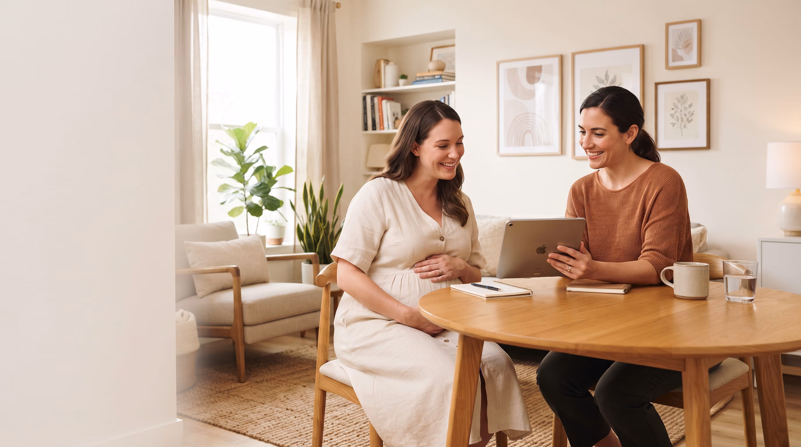 Pregnant woman smiling while consulting with a healthcare professional using a tablet at a wooden table.
