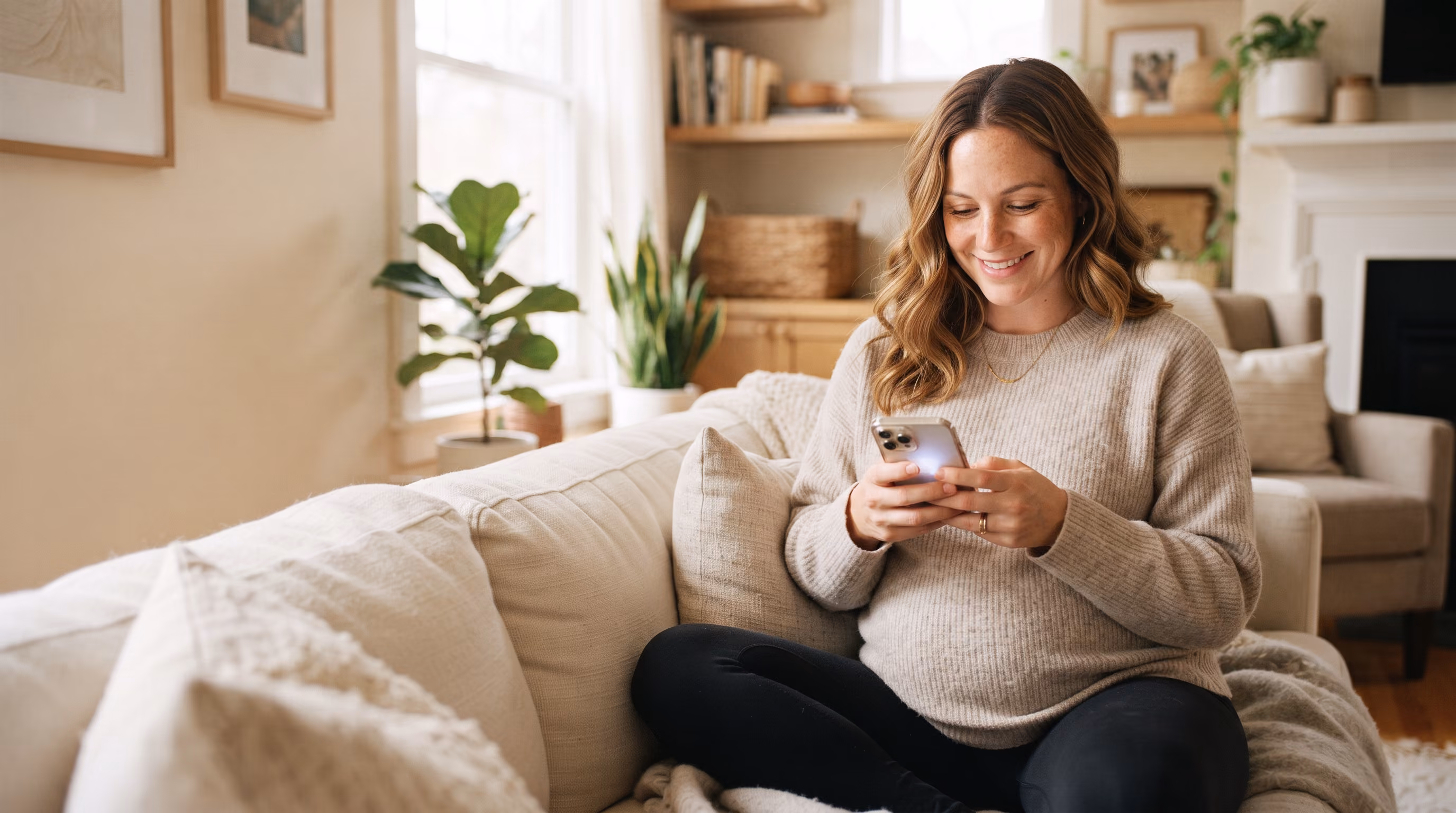 Pregnant woman sitting on a beige couch, smiling while using her smartphone in a cozy living room.