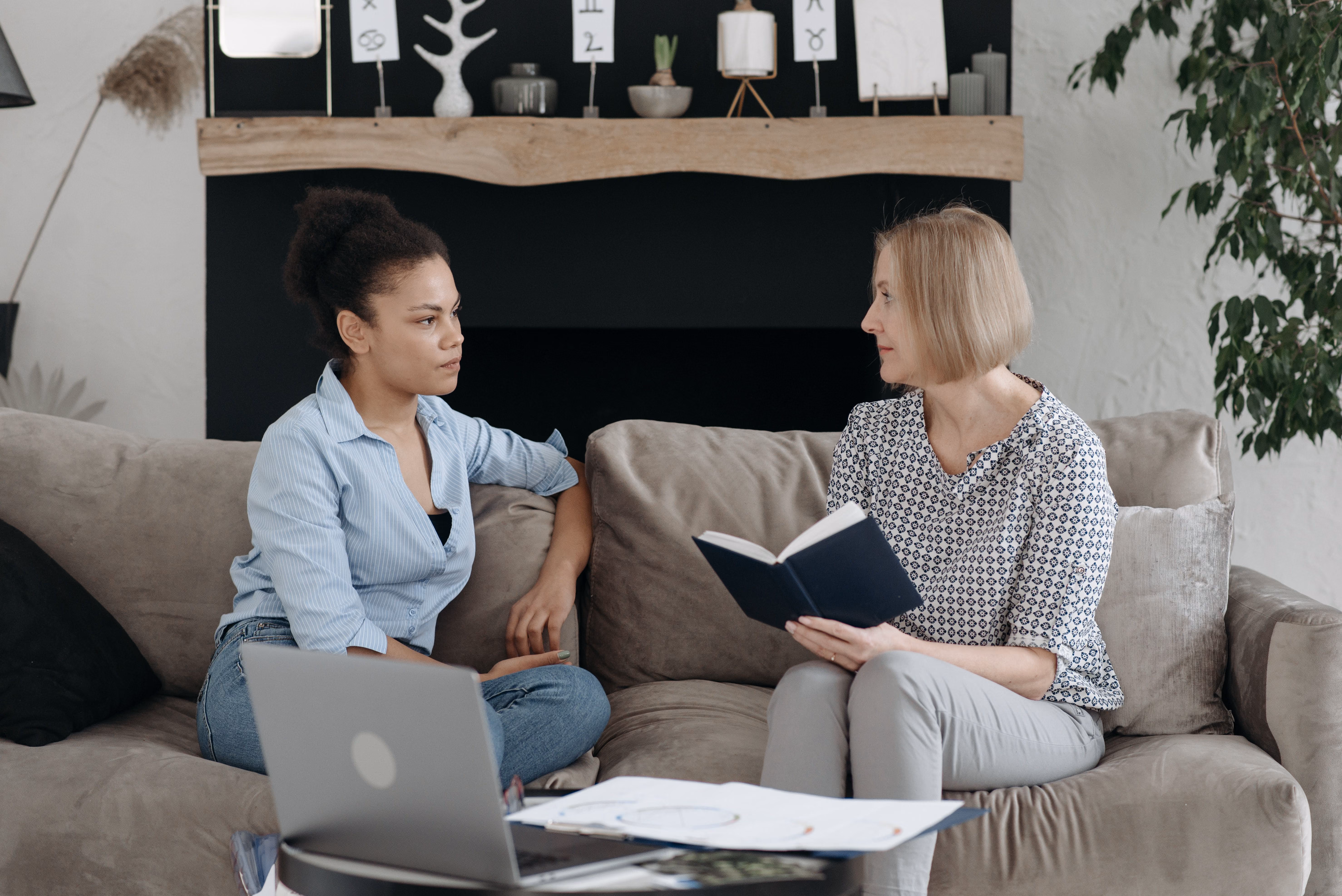 Two women sitting on a couch, one holding a book and the other listening attentively.