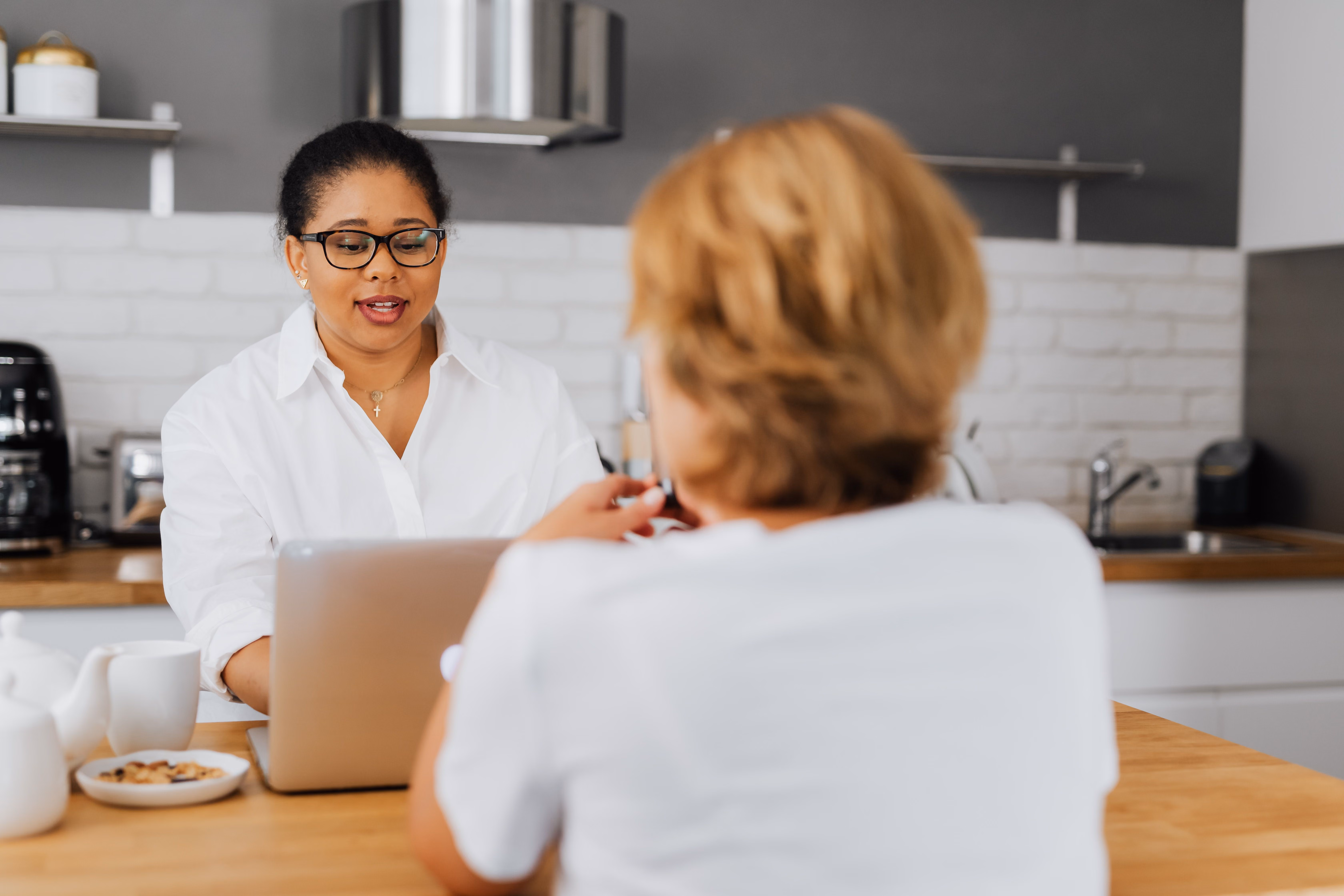 Two women sitting at a kitchen table, one using a laptop and talking to the other.