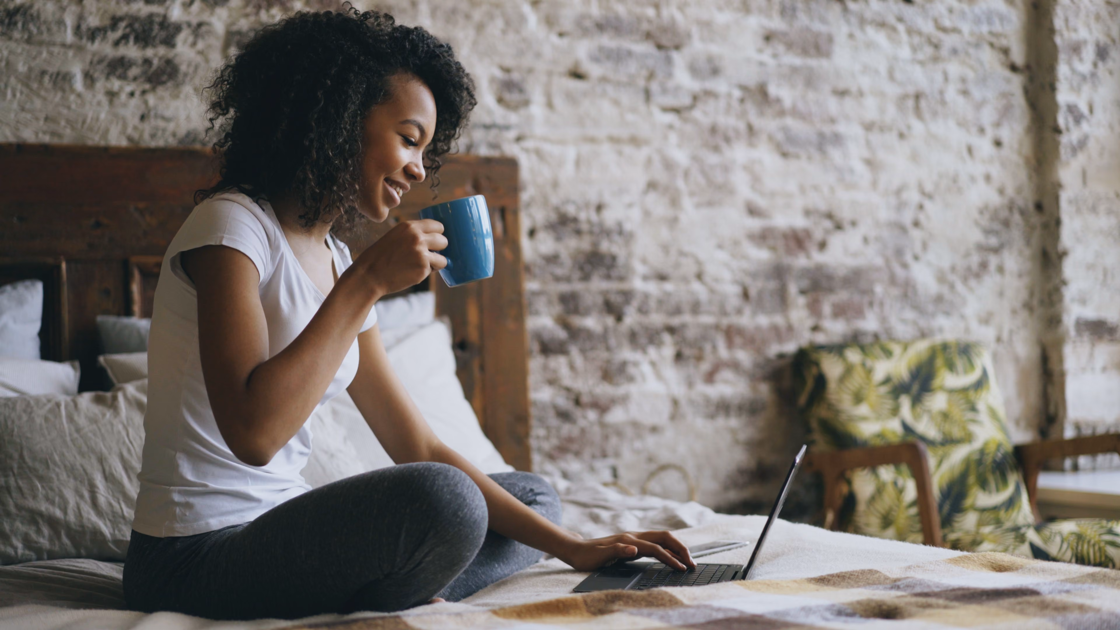 Smiling woman sitting cross-legged on a bed, holding a blue mug and using a laptop.