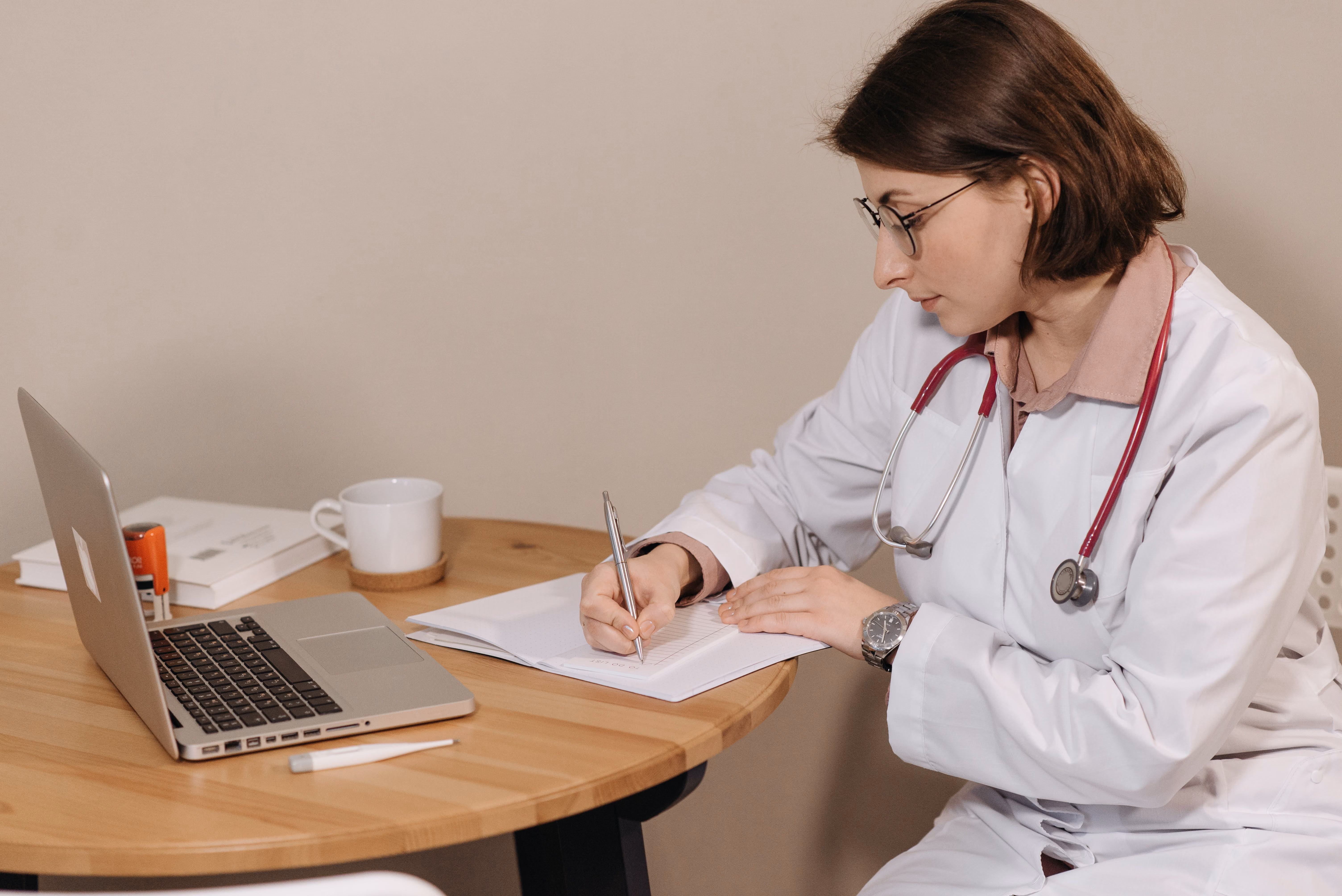 Female doctor in white coat writing notes at a wooden table with a laptop and a cup nearby.