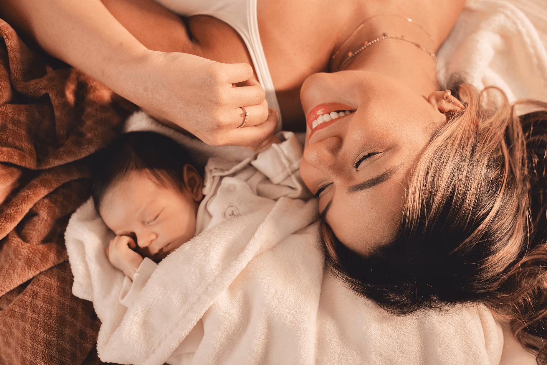 Smiling woman lying down next to a sleeping baby wrapped in a white blanket and resting on a brown blanket.