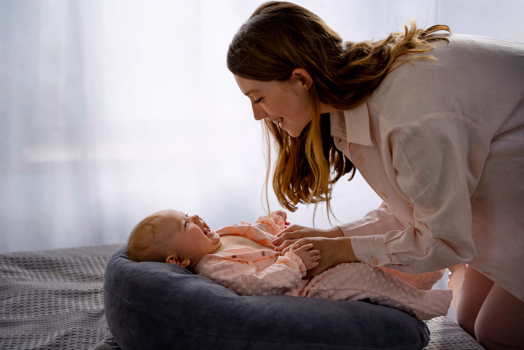A woman smiling and leaning over a baby who is lying in a dark cushioned baby lounger, both looking happy and engaged.
