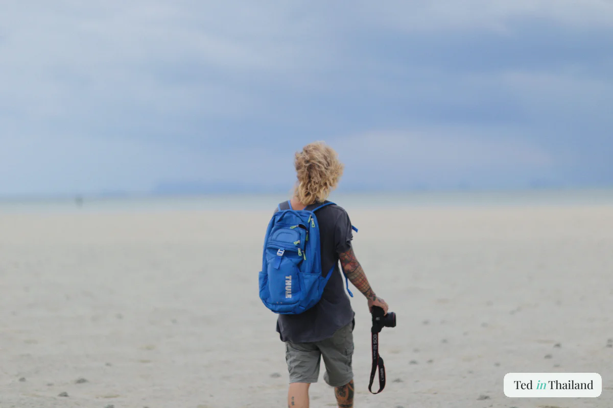 Solo backpacker with a blue backpack walking the wide pale sand of a Koh Samui beach on a budget trip