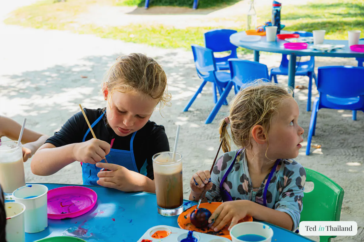 Two young girls painting and sipping iced drinks at a kids' club craft table on the sand at a Koh Samui resort