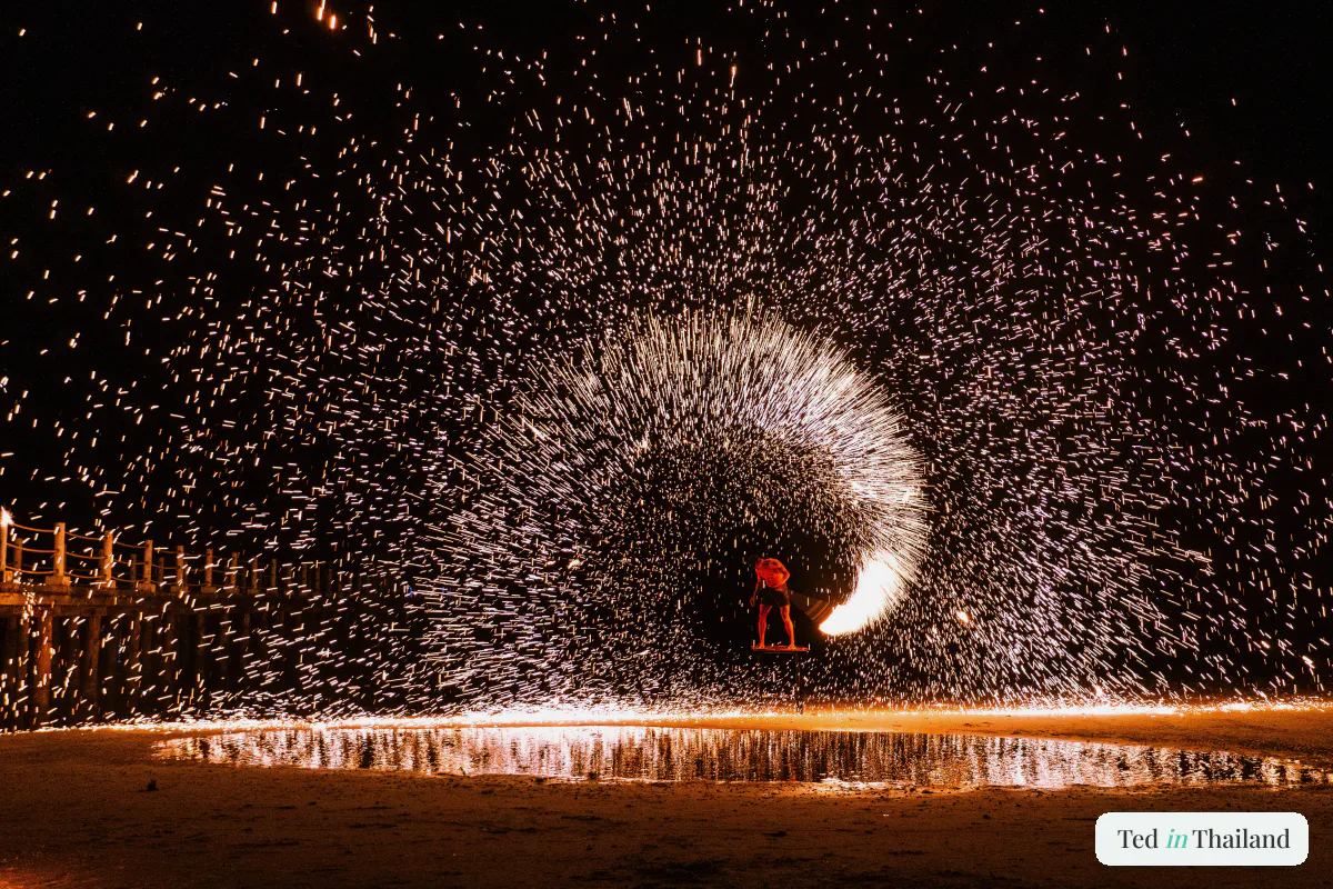 A fire performer spinning steel wool on a Koh Samui beach at night, a halo of sparks arcing across the dark sand