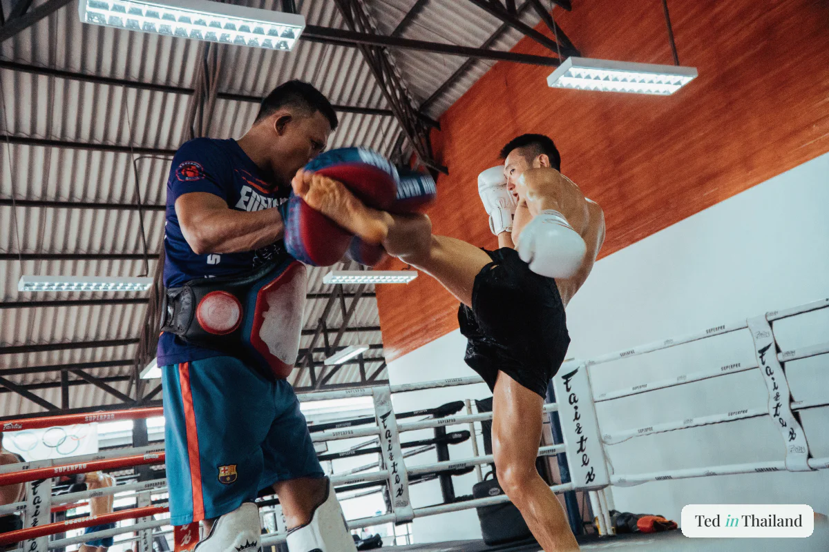 A Muay Thai trainer holding pads as a fighter lands a high kick during a training session in a Koh Samui gym