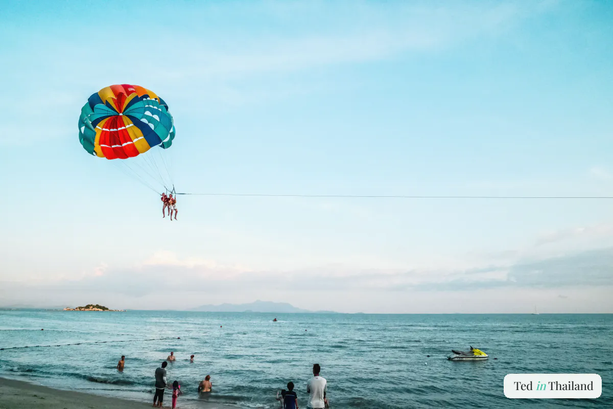 A colourful parasail carrying a rider high over the sea off Chaweng Beach on Koh Samui, swimmers and a jet ski in the shallows below