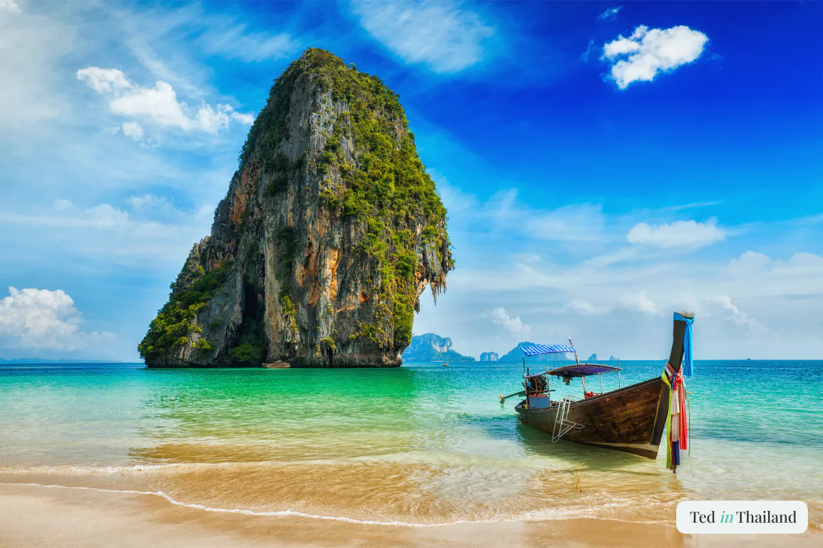 A traditional wooden long-tail boat resting on white sand beside clear water and a limestone island near Koh Samui