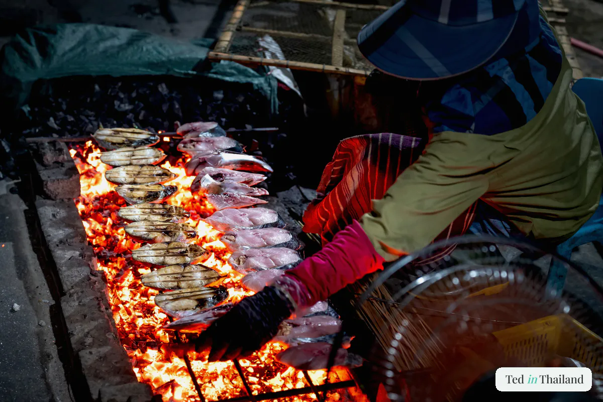 A street vendor grilling fish over glowing coals at a night market along Chaweng on Koh Samui