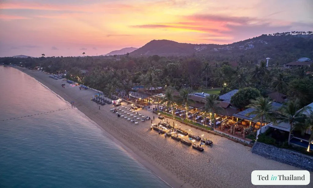 Aerial view at sunset of the lantern-lit beachfront restaurant and dining tables on the sand at Bandara Resort and Spa