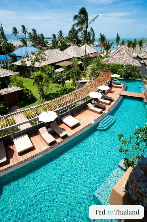 Winding turquoise lagoon pool and timber decking threading between Thai-roofed villas at Deva Beach Resort Samui