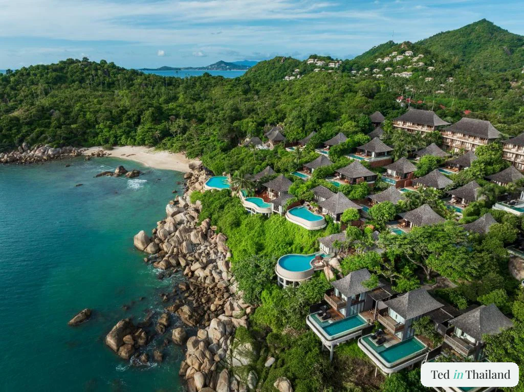 Aerial view of shingle-roofed pool villas stacked down a boulder headland above a private beach at Silavadee Pool Spa Resort