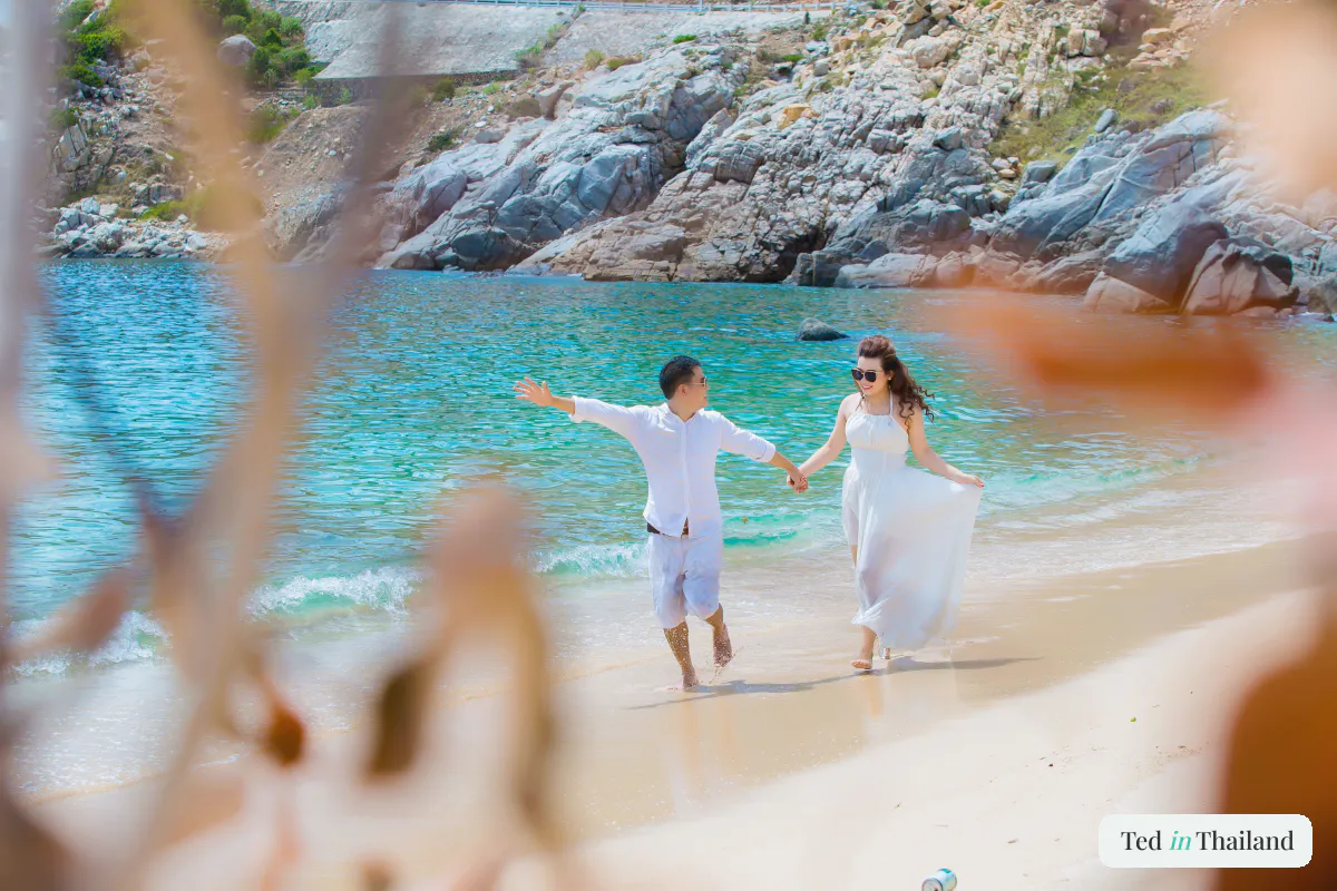 Couple in white walking hand in hand along a quiet cove beach during a honeymoon on Koh Samui