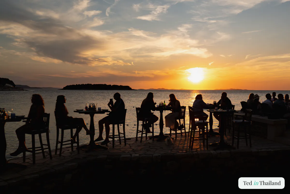 Diners at waterfront tables silhouetted against an orange sunset at a beachfront restaurant on Koh Samui