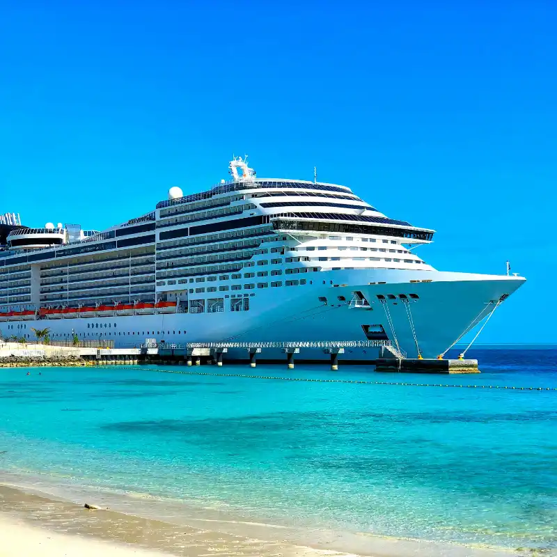 Large white luxury cruise ship docked by a turquoise sea and sandy beach under a clear blue sky.