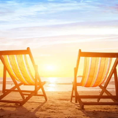Two empty striped beach chairs facing a glowing sunset over the ocean on sandy shore.