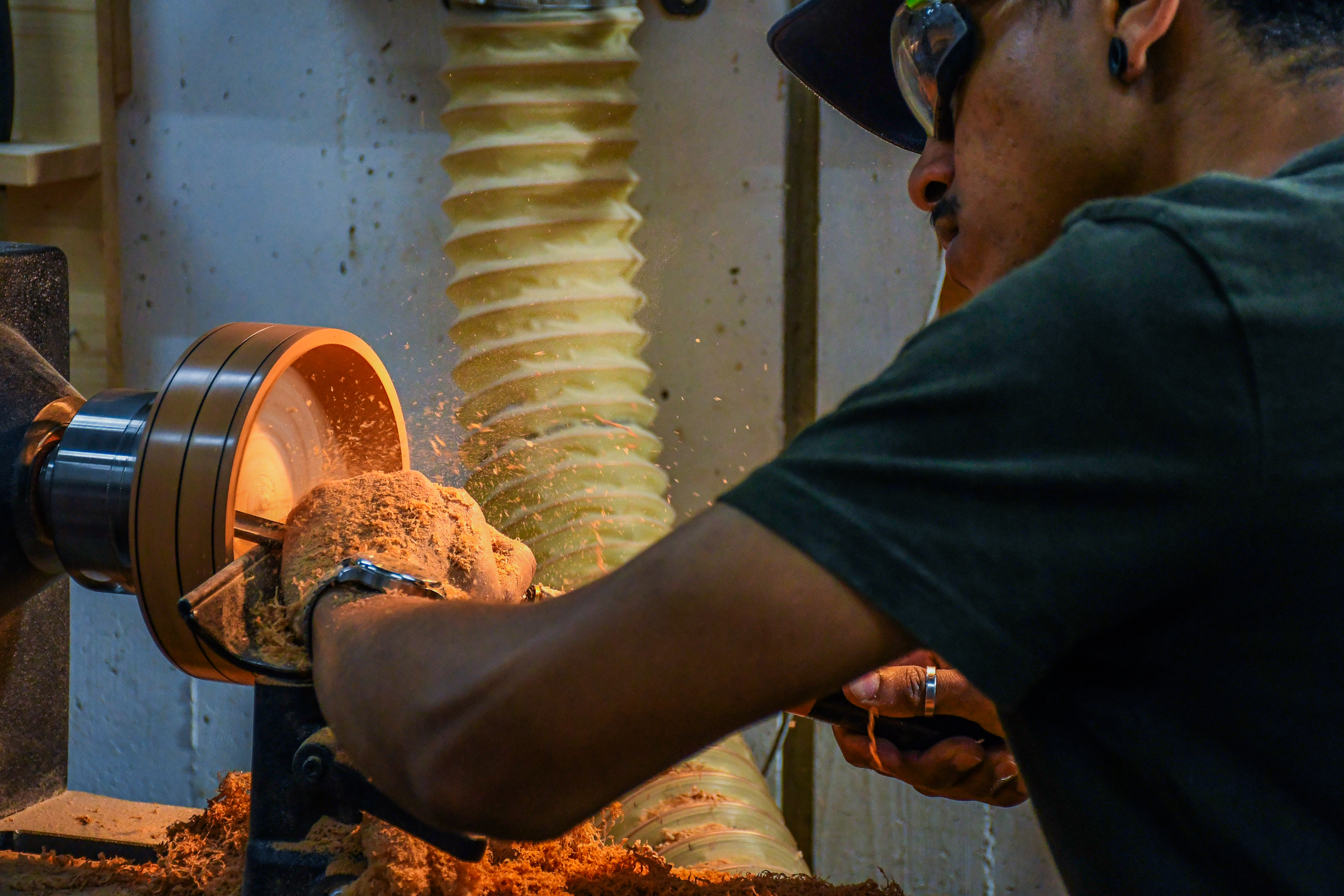 Person wearing safety glasses shaping a spinning piece of wood on a lathe with wood shavings flying.