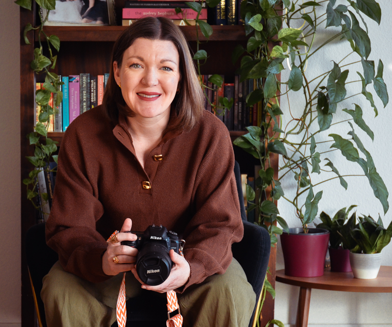 Smiling woman with brown hair sitting in front of a bookshelf and plants, holding a Nikon camera.