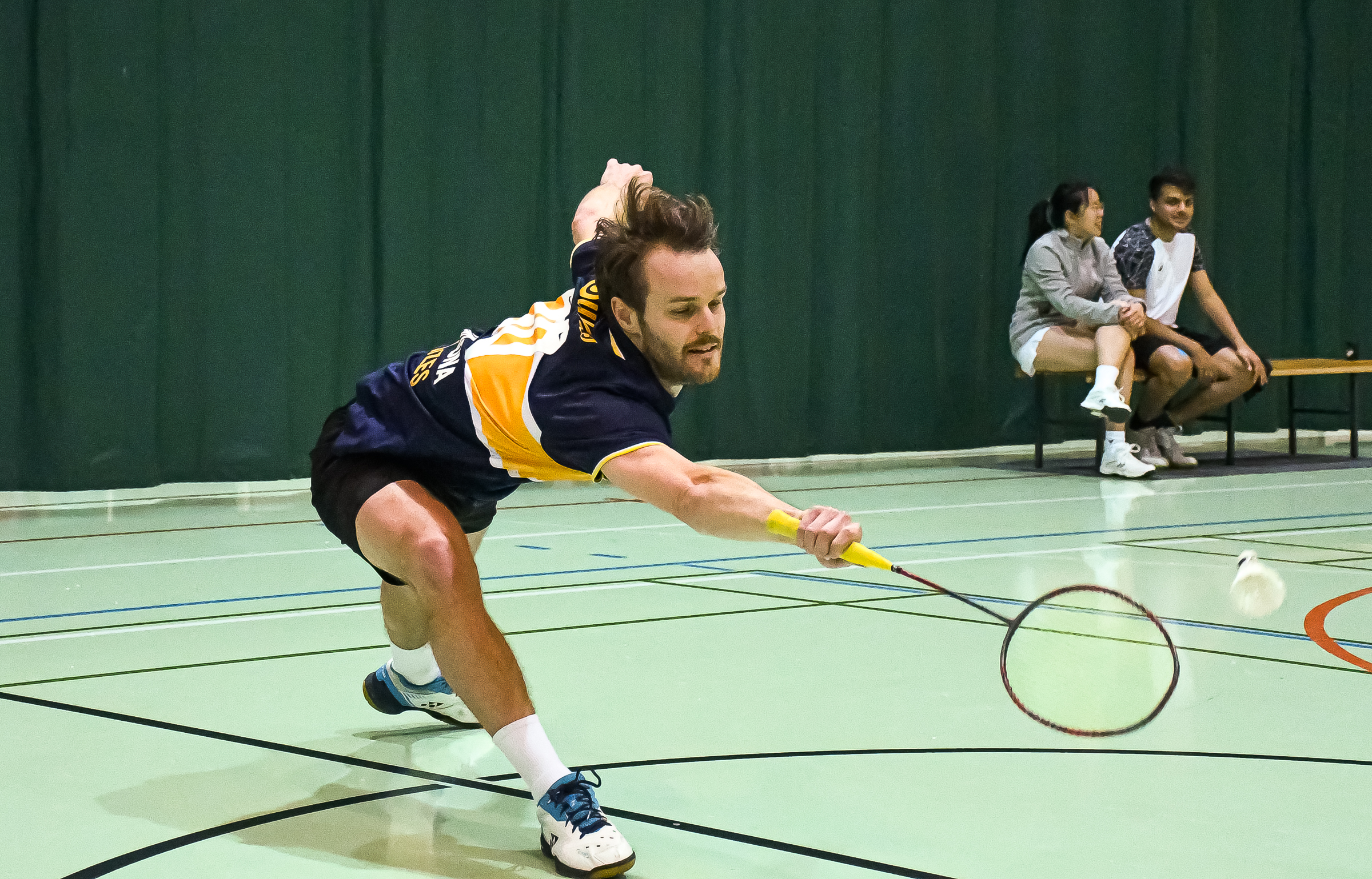 Male badminton player lunging forward to hit a shuttlecock with his racket in an indoor court.