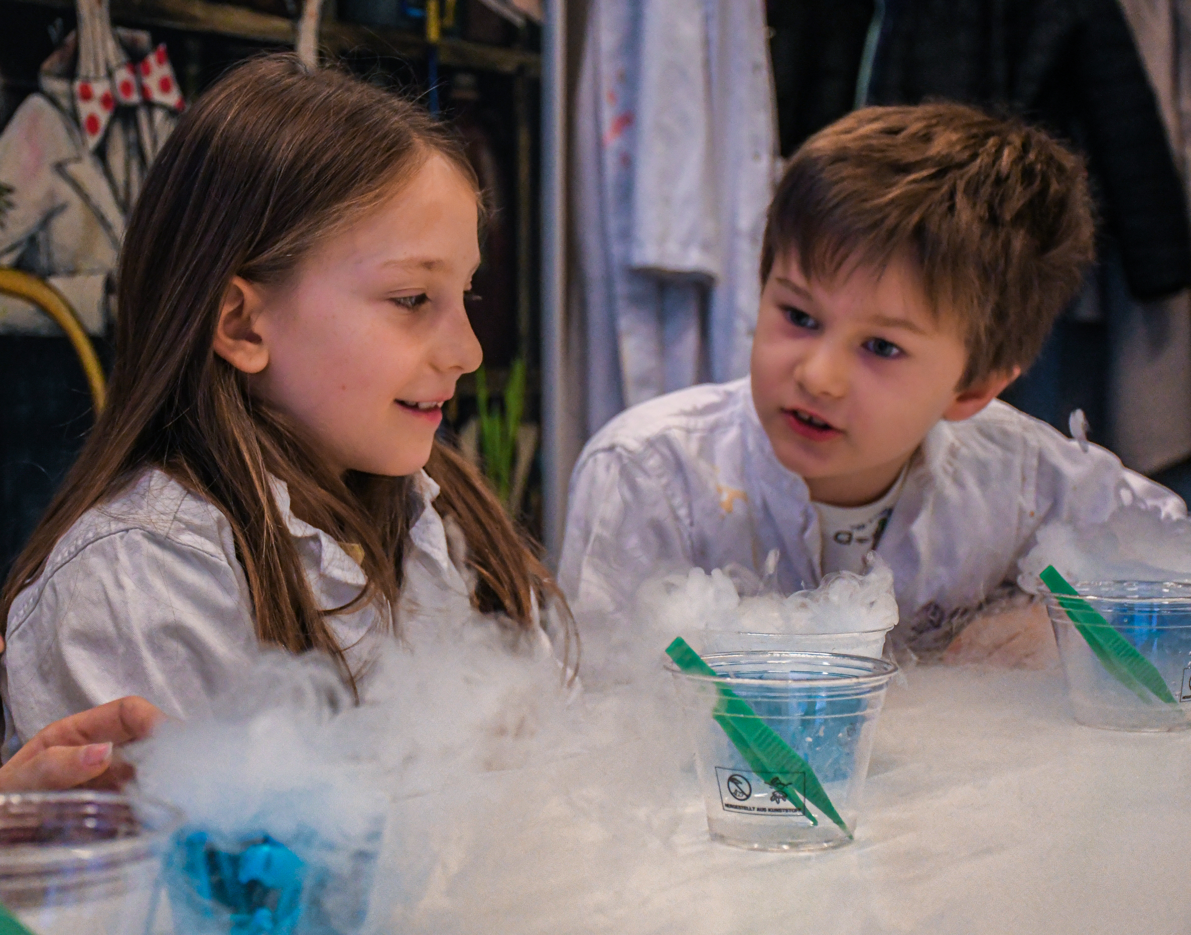 Two children wearing white lab coats observing a science experiment with smoke coming from clear plastic cups on a table.