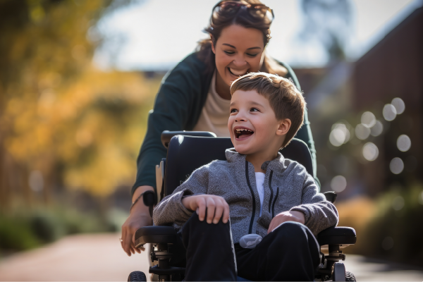 Woman smiling and pushing a laughing young boy in a wheelchair outdoors on a sunny day.