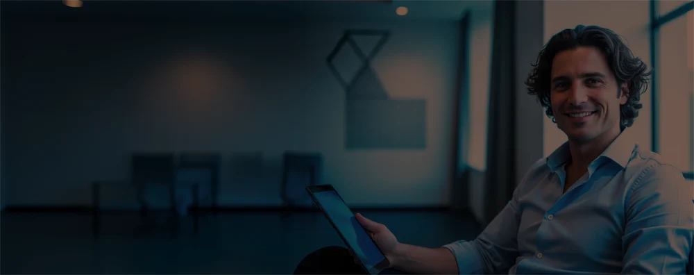 Smiling man in a blue shirt holding a tablet in a modern office with large windows.