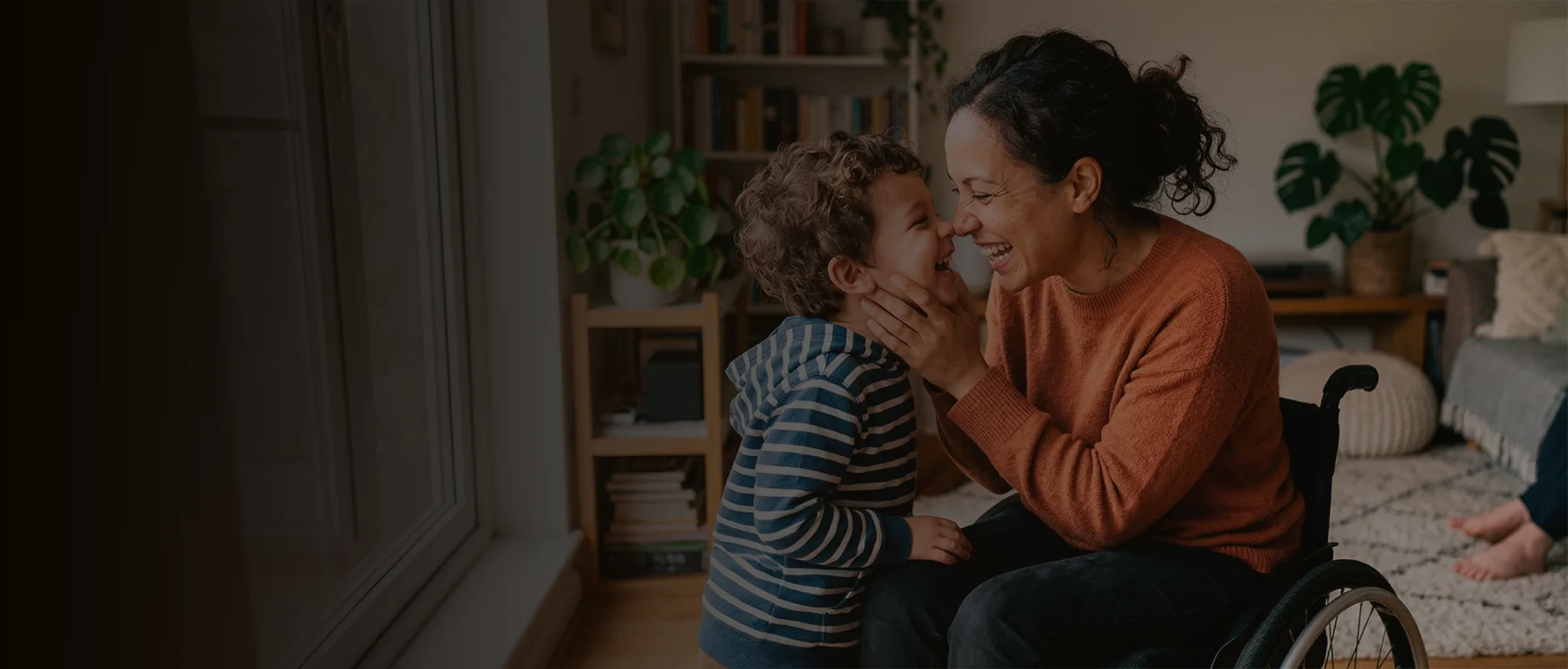 Woman in wheelchair smiling and touching noses with a laughing young child in a cozy living room.