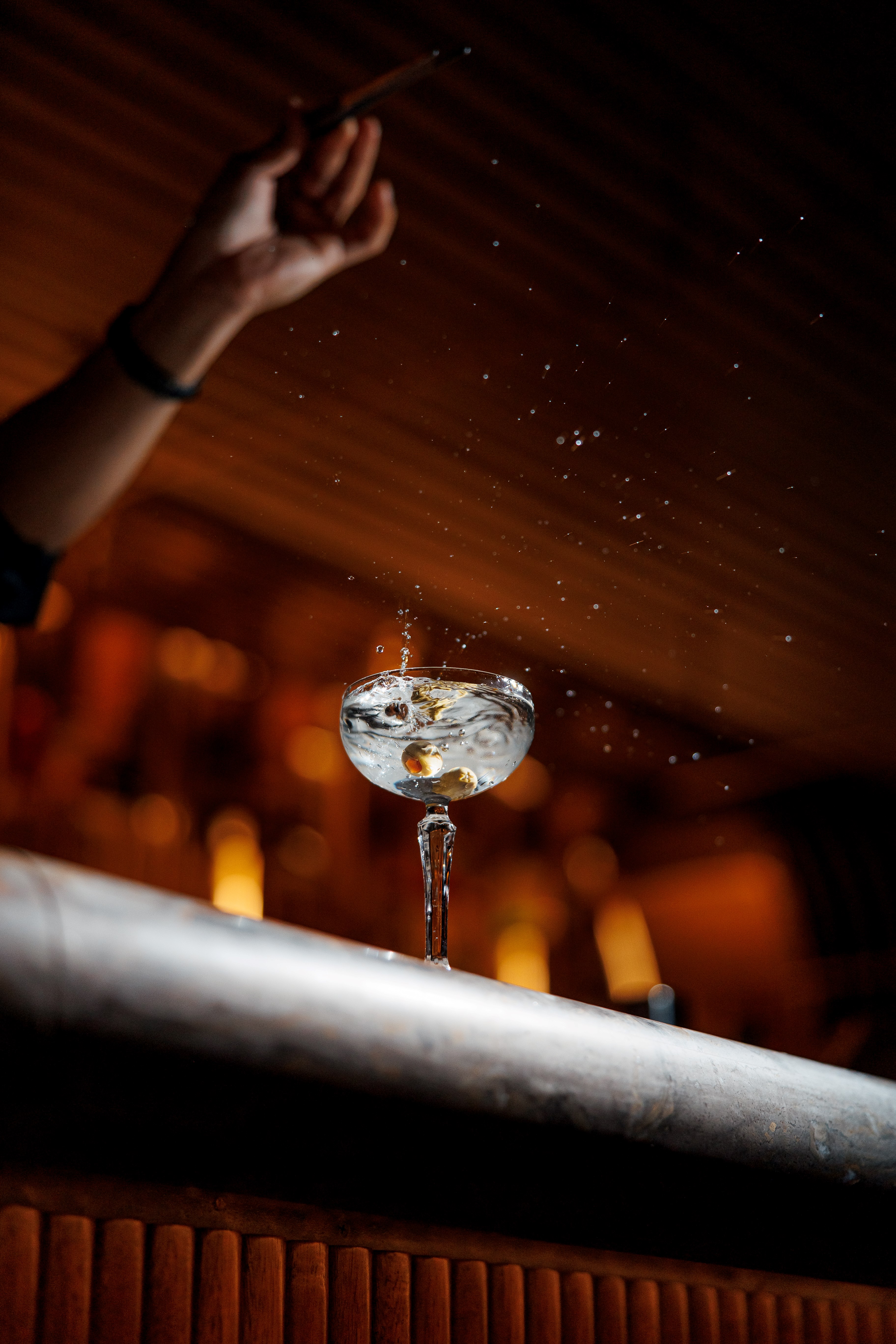 Close-up of a cocktail glass with olives splashing in liquid on a bar counter under dim warm light.