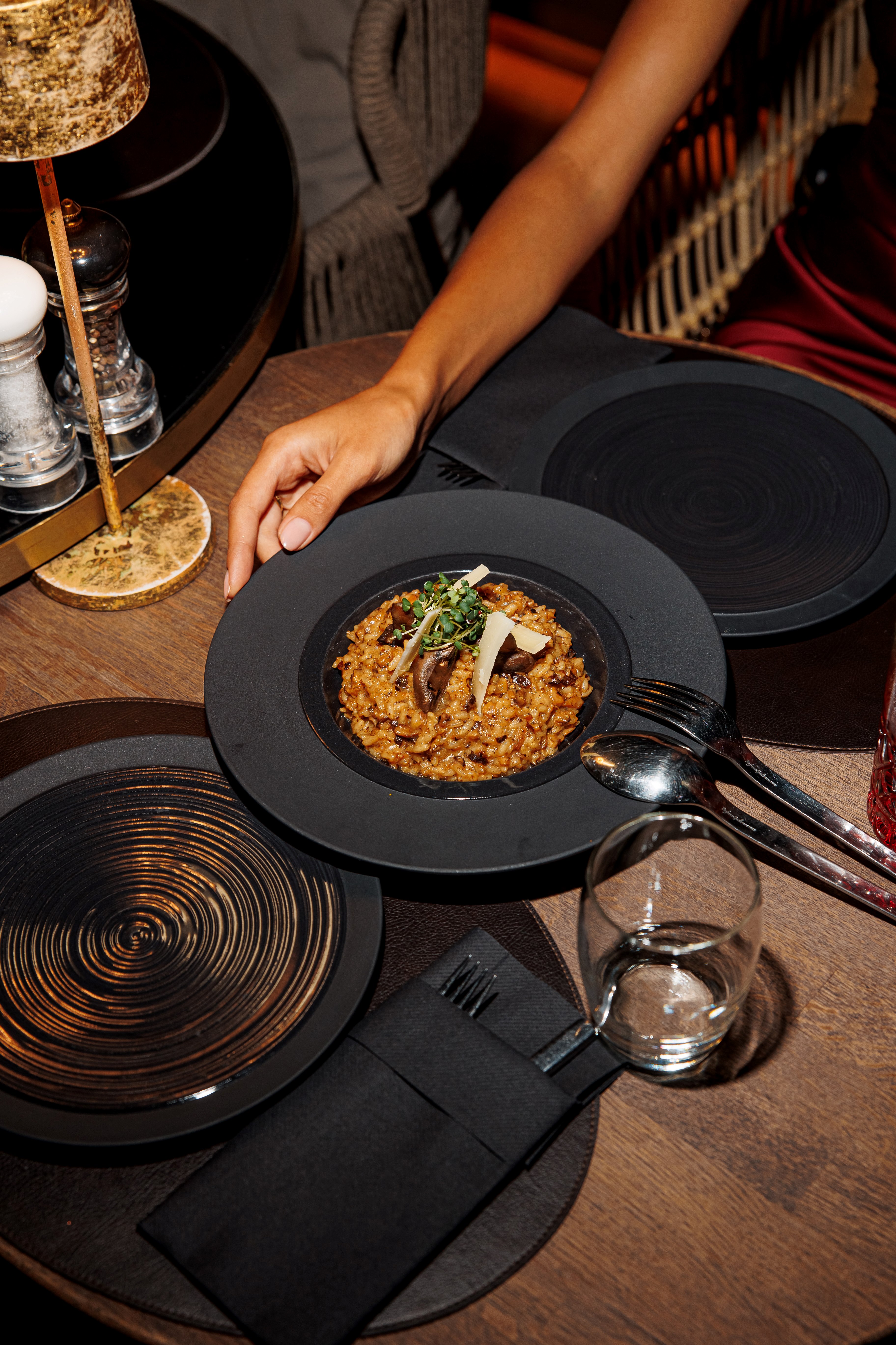 Hand holding black plate with mushroom risotto garnished with microgreens and cheese shavings.