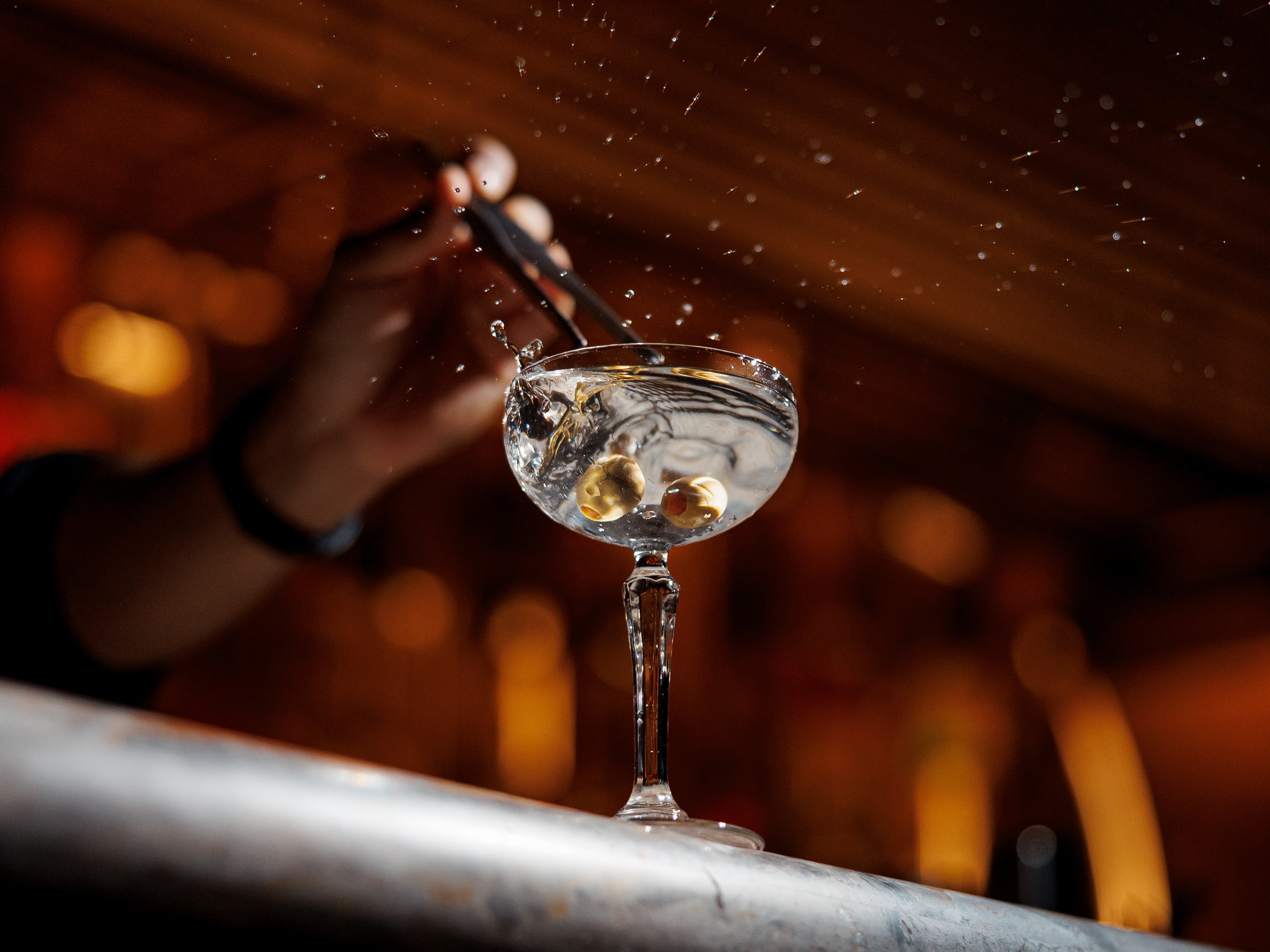 Clear cocktail in stemmed glass with olives splashing as hand drops them with tongs in dim bar light.