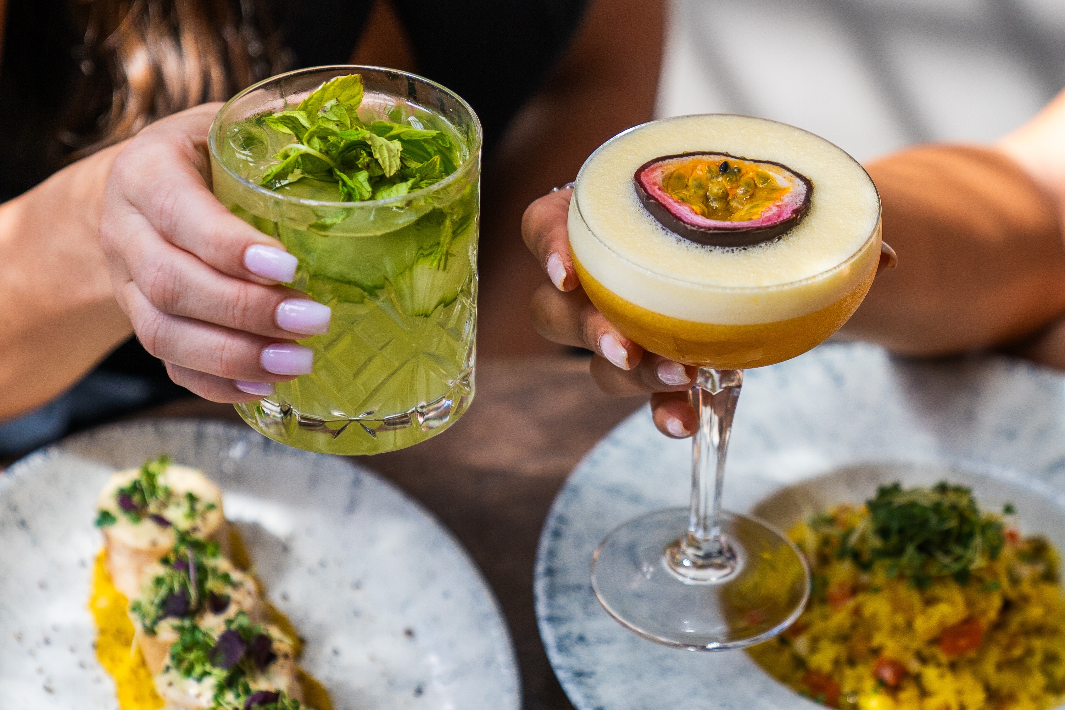 Two hands holding cocktails: a green herb drink and a passion fruit garnish drink with food plates below.